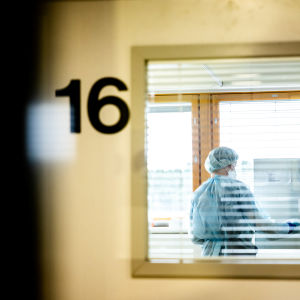 Sairaanhoitja koronapotilaiden eristyshuoneessa kuvattuna oven lasin läpi.