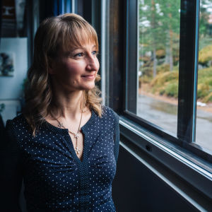 Tanja Poutiainen står i blå skjorta med vita prickar och tittar ut genom ett fönster.