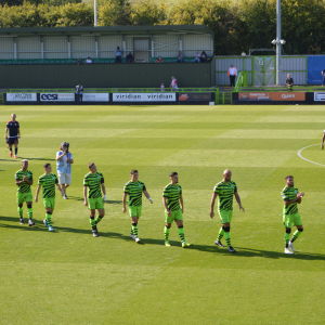 En fotbollsplan som det går människor på. I förgrunden går 10 män i en rad, de är klädda i gröna shorts och tröjor med svarta och gröna tröjor.