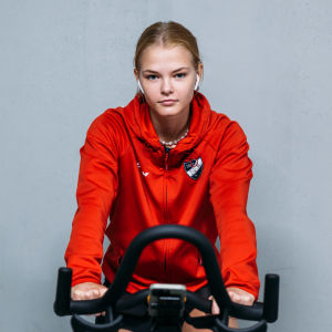 Nellie Karlberg istuu kuntopyörän selässä ja katsoo kameraan. Hän on vaalea, siro, 18-vuotias nainen, jolla on iPod-kuulokkeet korvillaan ja yllään HIFK-joukkueen paita.