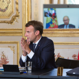 Presidentit Macron ja Putin keskustelevat videoyhteyden välityksellä. Putin istuu työpöytänsä äärelle, taustalla Putinin kasvot erottuvat televiosruudulta. 