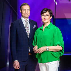 Mies sinisessä puvussa ja nainen vihreässä paidassa seisovat ja katsovat kameraan TV-studiossa.