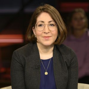 Deborah Feldman i brun page, stora glasögon med tunna bågar. Bruna ögon, svart kavaj och ett guldhalsband. Porträttfotografi.