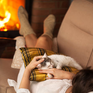 En kvinna som ligger i en soffa med en katt i famnen och en brasa i en kakelugn i bakgrunden.