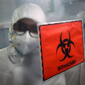 En person i skyddsoverall och munskydd står bakom en varningsskylt där det står "biohazard".