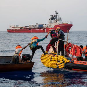 Migranter räddas från rostig fiskebåt till orange gummibåt på Medelhavet.