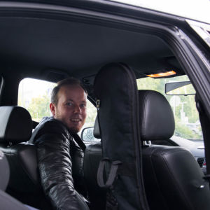 Mikael Grönroos har skrivit över 200 barnlåtar för Svenska Yles barnredaktion. Mikael Grönroos i bilen bakom ratten tittar över axeln mot kameran genom bakdörren som är öppen.