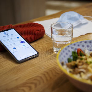 En mobil med Coronablinkern på skärmen ligger lutad mot en mössa på ett bord. Framför den en portion med nudlar, ett vattenglas och ett munskydd.