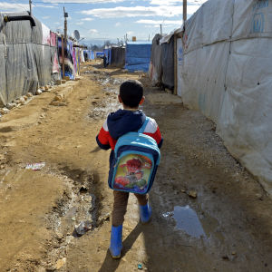 Ett syriskt flyktingbarn med blå ryggsäck promenerar i ett flyktingläger med tält i Libanon mit skolan.
