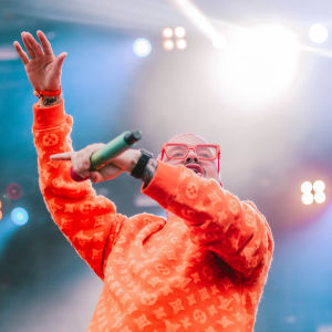 Oranssiin paitaan ja punaisiin aurinkolaseihin pukeutunut J Balvin kädet ilmassa Ruisrockin Niittylavalla. Kuva rajattu vyötäröstä alaspäin.