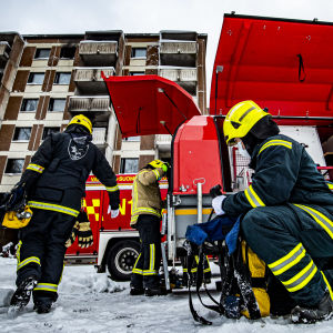Rökdykningsutrustning, lufttuber, brandslangar, munstycken, osv. Var och en har sin bestämda uppgift då brandkåren kommer fram till en höghusbrand. Tre brandmän plockar fram rökdykningsutrustning, brandslangar och munstycken ur brandbilar framför ett höghus, där många fönster och balkonger är svarta av eld och sot.