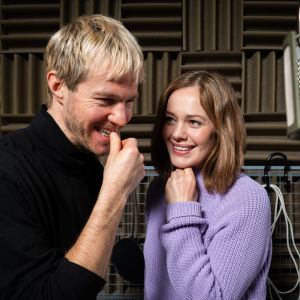 Kuvassa hymyilevä mies- ja naisnäyttelijä seisovat kuunnelmastudiossa katosta roikkuvan mikrofonin vieressä.