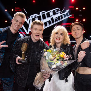 Tre unga män och en ung kvinna ler och tittar in i kameran efter finalen i The Voice of Finland. Vinnaren har en gyllene mikrofon och en blombukett i handen,