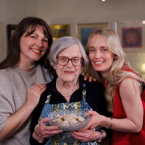 Kolme naista hymyilee ja keskimmäisellä vanhalla naisella on kädessään lasikulho, jossa on lusikkaleipäsiä.
