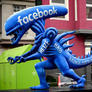 Sinisen alien patsaan hännästä pitää kiinni lennossa oleva tuomarihahmo. Alien edustaa Facebookkia ja tuomari oikeutta.