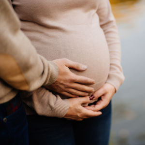 En gravid kvinna med tröja på som håller händerna runt magen. En annan person står bredvid och håller också ena handen runt magen.