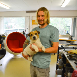 En man som håller en hund i famnen. I bakgrunden stolar.