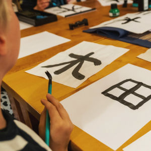 Pöydällä papereita, joissa japanilaisia kirjoitusmerkkejä, sivustalla näkyy lasta, jonka kädessä sivellin