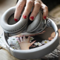 En hand med rött slitet nagellack håller i ett par hörlurar som vilar på en tidning med en kvinna på omslaget.