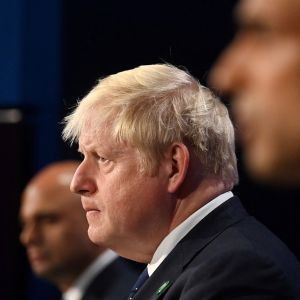 Boris Johnsonin kasvot tarkennettuna kuvassa. Molemmilla puolilla istuu ihminen, jonka kasvot ovat sumeat kuvassa. Kolme ihmistä istuu rivissä ja katsoo kuvassa vasemmalle.