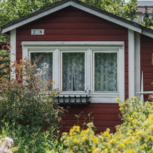 Punainen tupa valkoisin ikkunanpuittein vehreässä puutarhassa. Taustalla moderni kerrostalorakennus.