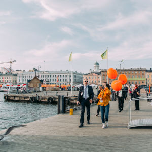 People walking on a dock. Three orange ballons in the foreground.