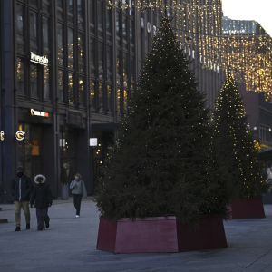Två granar med julljus står i förgrunden, på gatan går vinterklädda människor. På byggnaderna hänger julljus.