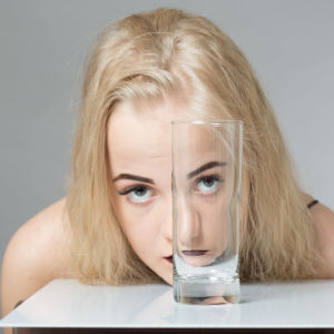 Blond ung kvinna tittar mot kameran genom ettt tomt dricksglas