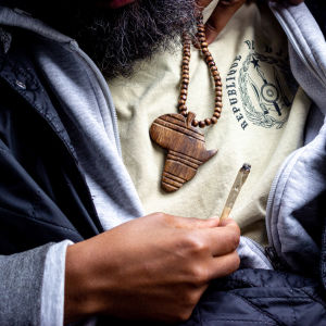 Närbild tagen på bröstkorgshöjd av en man med ett Afrika-halsband och en joint i handen.
