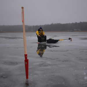 I förgrunden står en isbill med röd spets. En man i flytdräkt, en gul overall och blå mössa, sitter på en ryggsäck med pall och pimpelfiskar på en sjö där mycket smältvatten samlats på isen.
