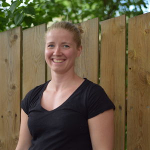 En blond kvinna i svart t-skjorta står vid ett träfärgat staket och ler mot kameran. Sommar.