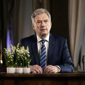 President Sauli Niinistö sitter vid ett skrivbord i kostym och talar. På bordet finns en mikrofon och blommor.