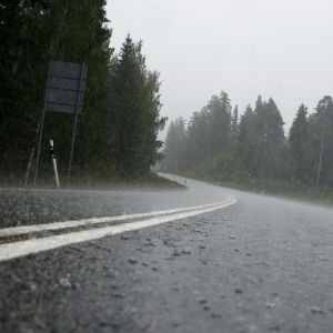 Metsän läpi kaartuva kaksi kaistainen autotie, jonka pinta on sateesta märkä ja roiskuu rankkasateessa.