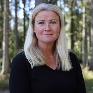 Kvinna med ljust hår och svart jumper står i en skogsglänta.