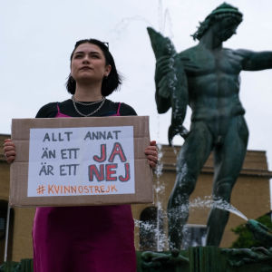 Ung kvinna håller i skylt med texten "Allt annat än ett JA är ett NEJ, #kvinnostrejk". I bakgrunden stor staty föreställande en naken grekisk gud.