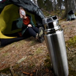 En kvinna sitter halvt inne i ett tält i skogen och för en mugg till munnen. I förgrunden en öppnad termos.