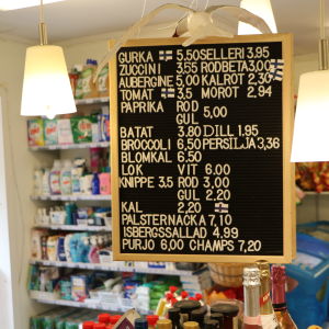 Bybutiken håller öppet året om. En prislista i en bybutik, med fulla hyllor av hushållsvaror i bakgrunden.