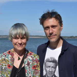 Konstnärerna Emma Fält och Roberto Fusco tittar och ler mot kameran. I bakgrunden havet, en färja och blå himmel.