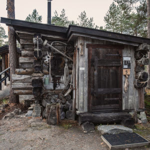 Pieni kelohongasta tehty mökki, jonka ulkoseiniä koristaa eläimen talja, sarvet ja työkaluja.
