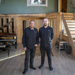 Två män i  en stor inspelningsstudio i två våningar med träklädda väggar och tak. I det stora rummet finns bland  annat flygel, gitarrer, matta på golvet