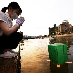 Henkilö kumartaa vesilyhdyn vieressä, taustalla näkyy Hiroshiman pommituksen muistomerkki.