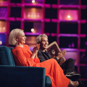 Nauravainen Krisse Salminen istuu sohvalla punaisessa mekossaan, vierellään Miika Nousiainen.