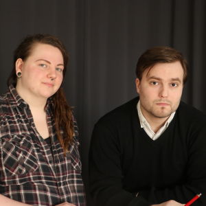 En ung kvinna med långt rödbrunt hår står bredvid en ung man som lutar sig mot ett bord.