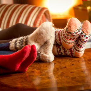 Tre personer sitter i soffan med fötterna i yllesockor som de vilar på bordet: En brasa och presentpaket finns i bakgrunden.