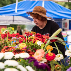 Kukkakuhhauksen myyntikojussa, kukkakimppujen takana, olkihattupäinen nainen järjestelee kukkasia.