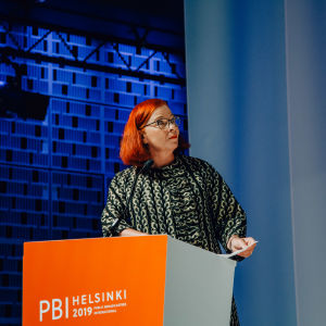 A woman behind an orange podium on stage, looking towards what seems to be a screen.