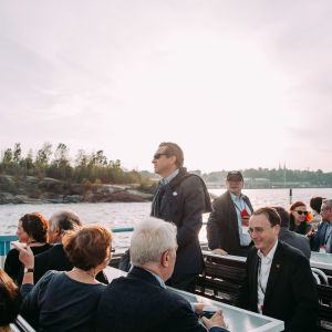 A man standing on an outside deck on a ferry. More people sitting down.