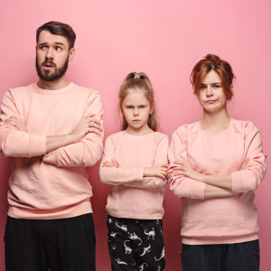 Pappa, mamma och dotter framför rosa fondvägg, alla med korsade armar och sura miner
