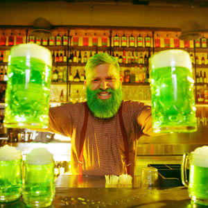 Har de ens grön öl på Irland? Bartender med grönt skägg och grönt hår håller fram två stop med ilsket grön öl och fler gröna ölsttop står på bardisken.
