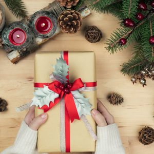 Händer håller i en julklapp paketerat i beige presentpapper och rött och silver band. På bordet finns också dekorerat granris, kottar och röda stearinljus.